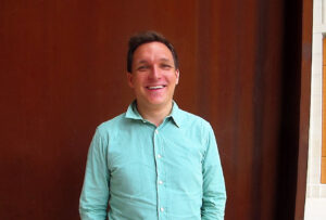 A man wearing a turquoise sweater stands smiling