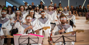 A crowd of people sit on roman steps wearing white robes and white sunglasses