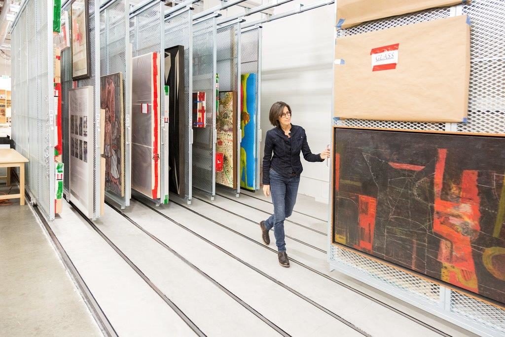 A woman stands by a rack of paintings in storage
