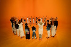 A group of people stand in a circle with arms upraised against an orange background, Holman