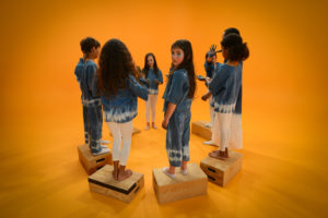 A group of children stand in a circle on apple boxes against an orange background, Holman
