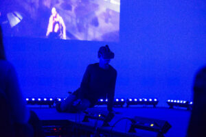 A man wearing a VR headset sits on a stage bathed in blue light with a video projection behind him as audience members look on