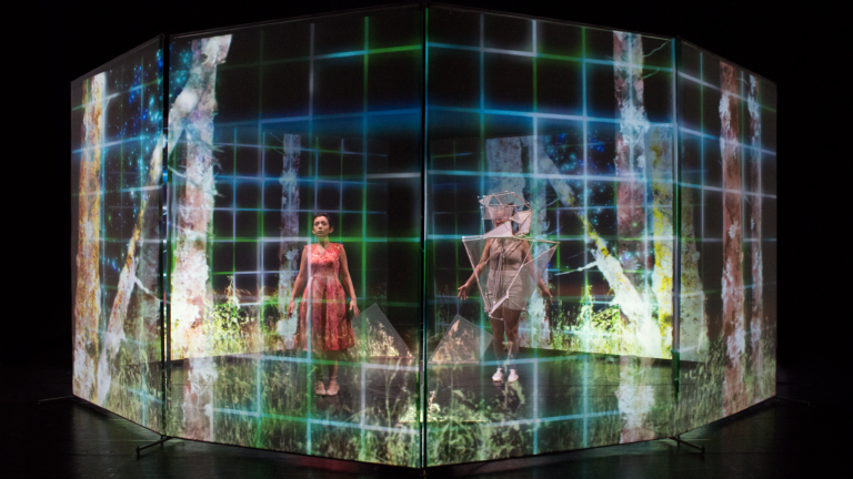 Two costumed performers stand behind an octagonal scrim with projected images