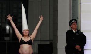 A Caucasian woman in a black bra wears a white dunce's cap and raises her arms
