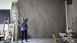 Film still of a woman painting on a large canvas, using a long paintbrush to reach the top.