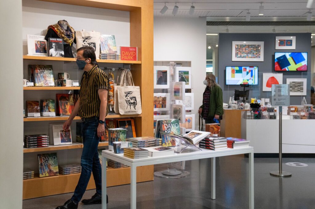 Two people wearing masks walking through a museum store with shelves of books and art prints.