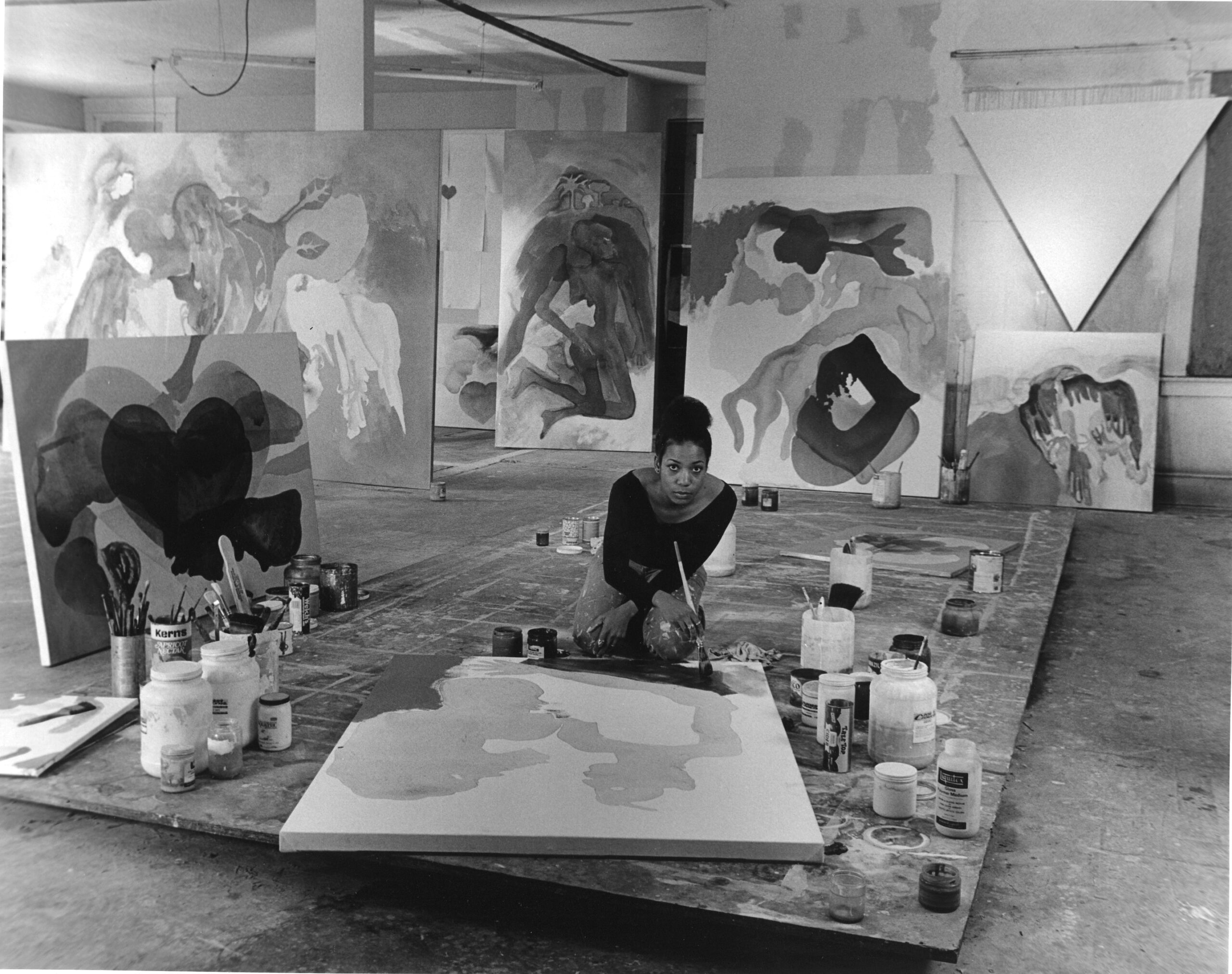 =A black and white photograph of artist Suzanne Jackson painting in her studio with large abstract artworks and paint supplies in the background.