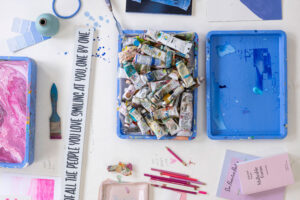 Creative artist workspace with paint tubes, brushes, color swatches, marbled tray, inspirational quote, and art supplies on white background.