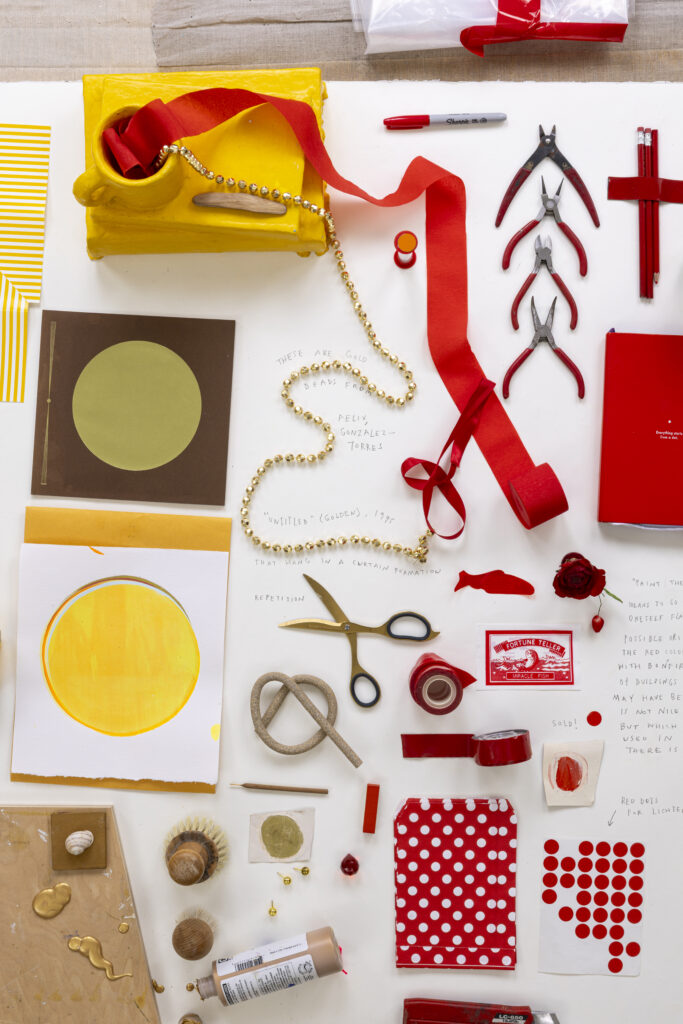 DIY craft supplies flat lay with red ribbon, beads, scissors, pliers, paint tubes, colored paper, and a yellow gift bag on white background.