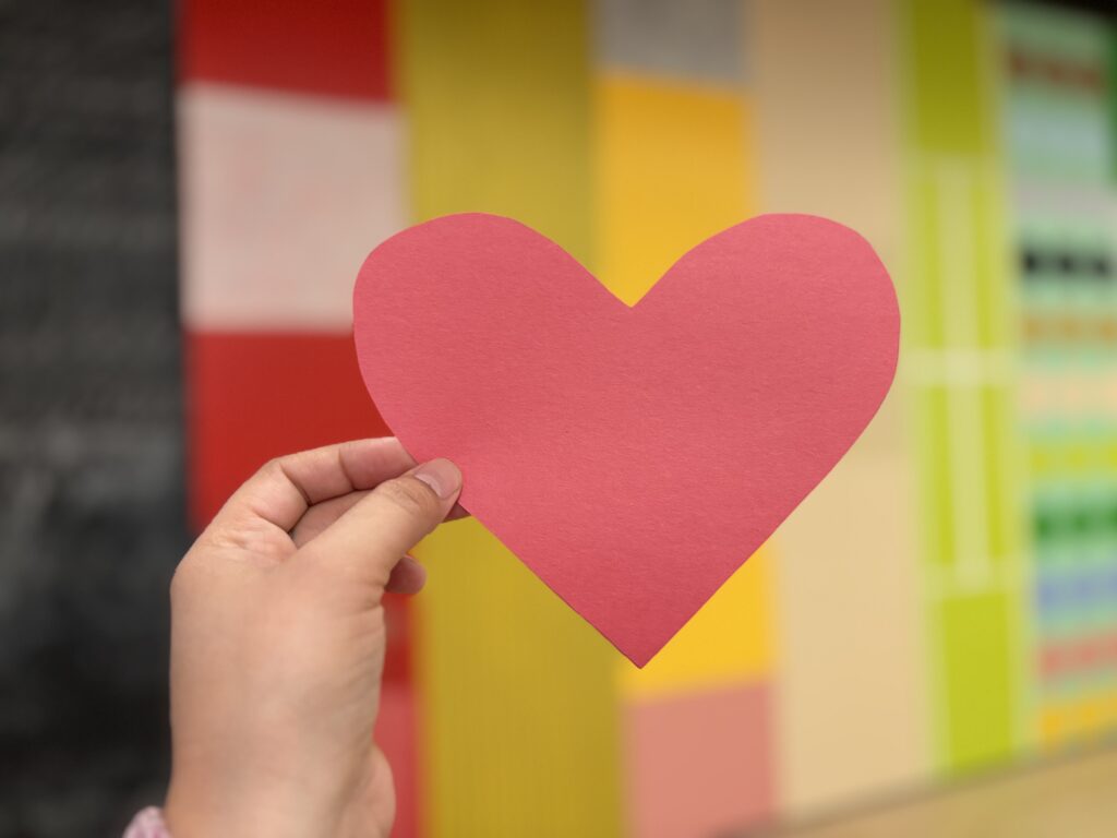 A hand holding a red paper heart in front of a colorful background.