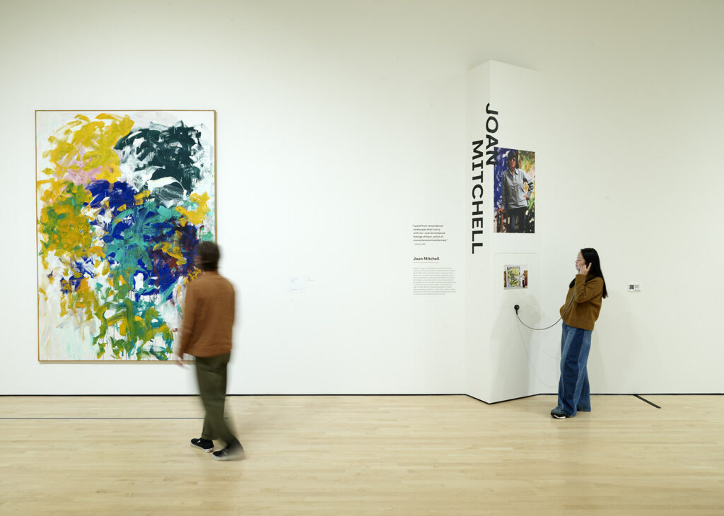 Two people in a museum gallery: one is walking toward a colorful abstract painting while the other is listening to an audio guide while standing below a sign that says "Joan Mitchell" in large black graphic letters.